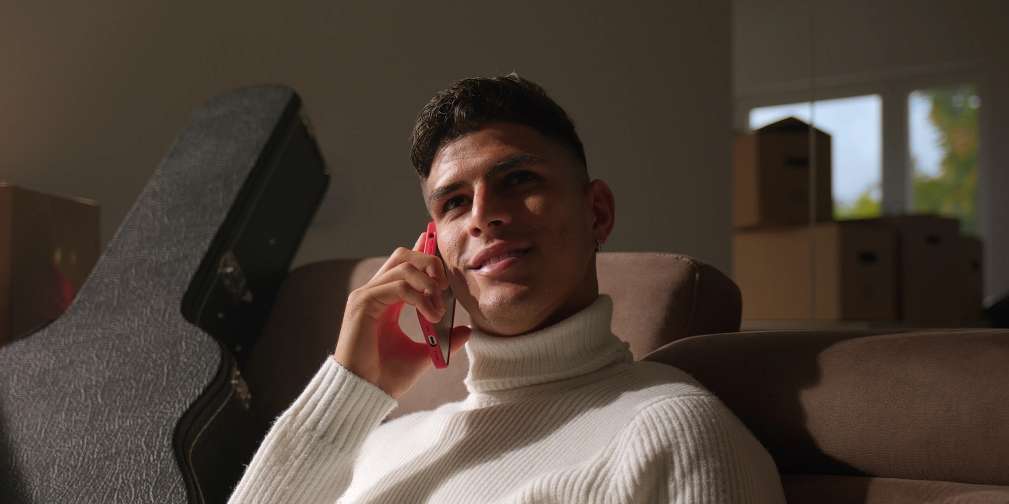 A young soccer player, Piero Hincapié, sits on his couch and makes a phone call. He is wearing a white sweater. Next to him is a guitar case, and moving boxes can be seen in the background.