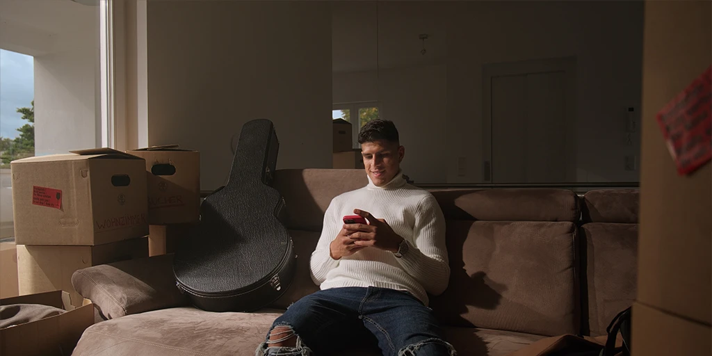 A young soccer player (Piero Hincapié) sits on his couch and taps on his cell phone. He is wearing a white sweater. The sun shines through the balcony door. Next to him are moving boxes.