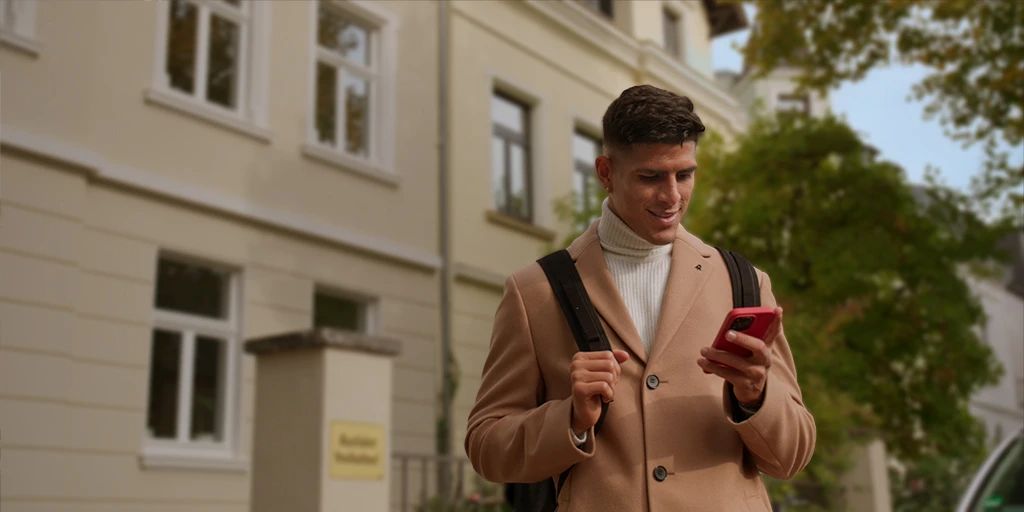 A young man (Piero Hincapié) wears a coat and looks at his cell phone. He is carrying a backpack and stands in front of a yellowish house.