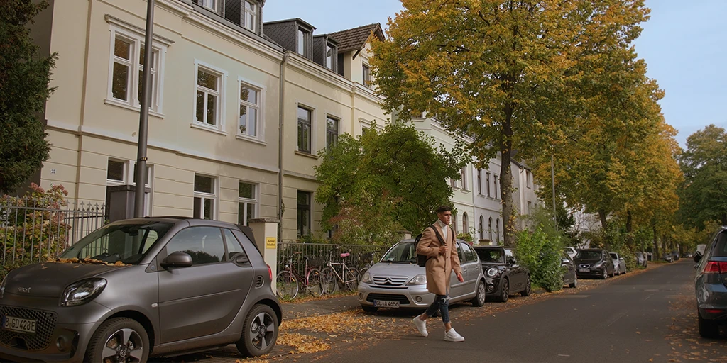 A young man (Piero Hincapié) crosses a street. He is wearing a brown coat. Behind him is a yellowish house. In the distance is an avenue with autumnal trees.