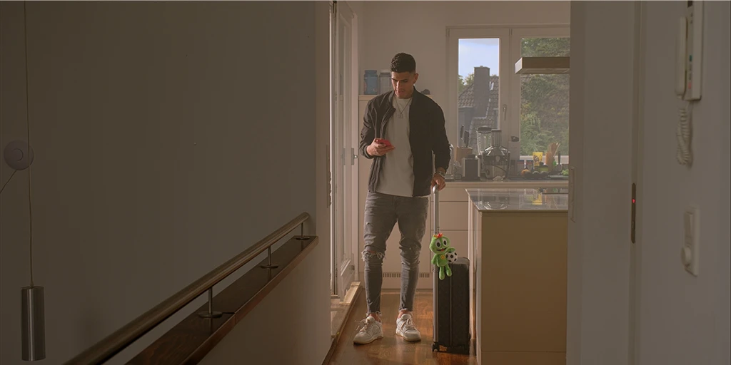 A young man (Piero Hincapié) stands in his kitchen and holds a cell phone in his hand. With his left hand, he holds a suitcase with a green mascot sitting on it. Sunlight hits the soccer player from the side.