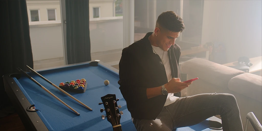 A young man (Piero Hincapié) sits in his villa on a blue pool table. He holds a cell phone in his hand. The room is flooded with sunlight. Next to him is a guitar.