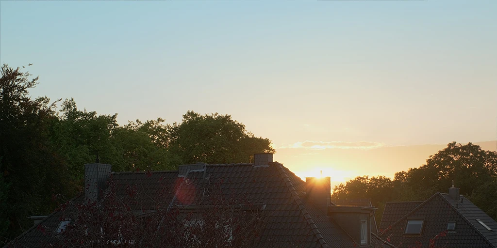 The sun sets and disappears behind a chimney of a residential house. Two roofs can be seen. Behind it is a forest. The sky shows a blue-red-yellow gradient.