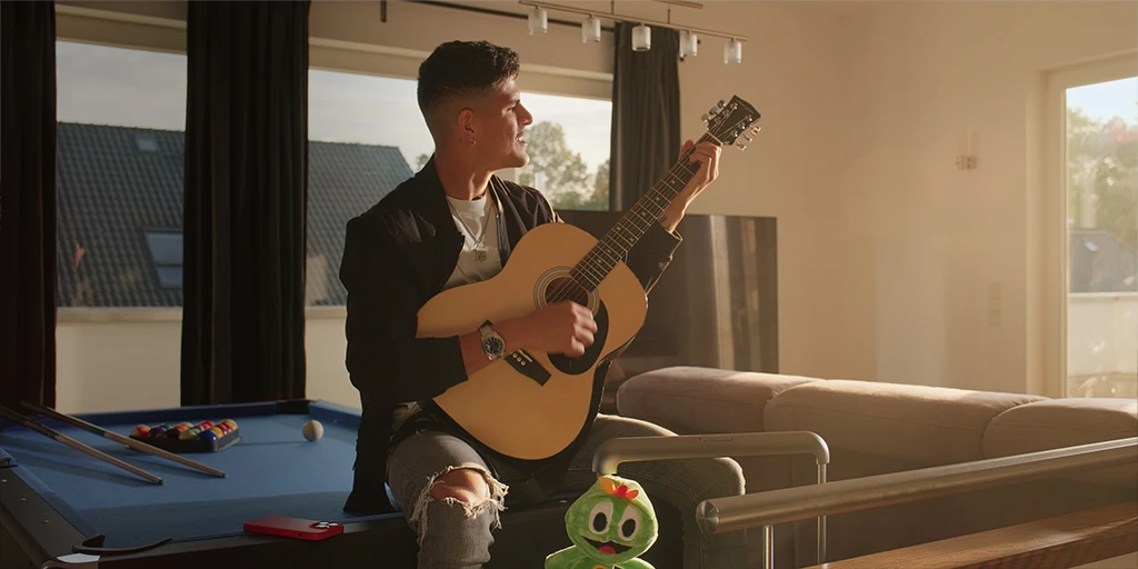 A man (Piero Hincapié) plays guitar and sits on his pool table. The room is flooded with sunlight, and a suitcase stands in front of Piero. A mascot looks cheerfully into the camera.