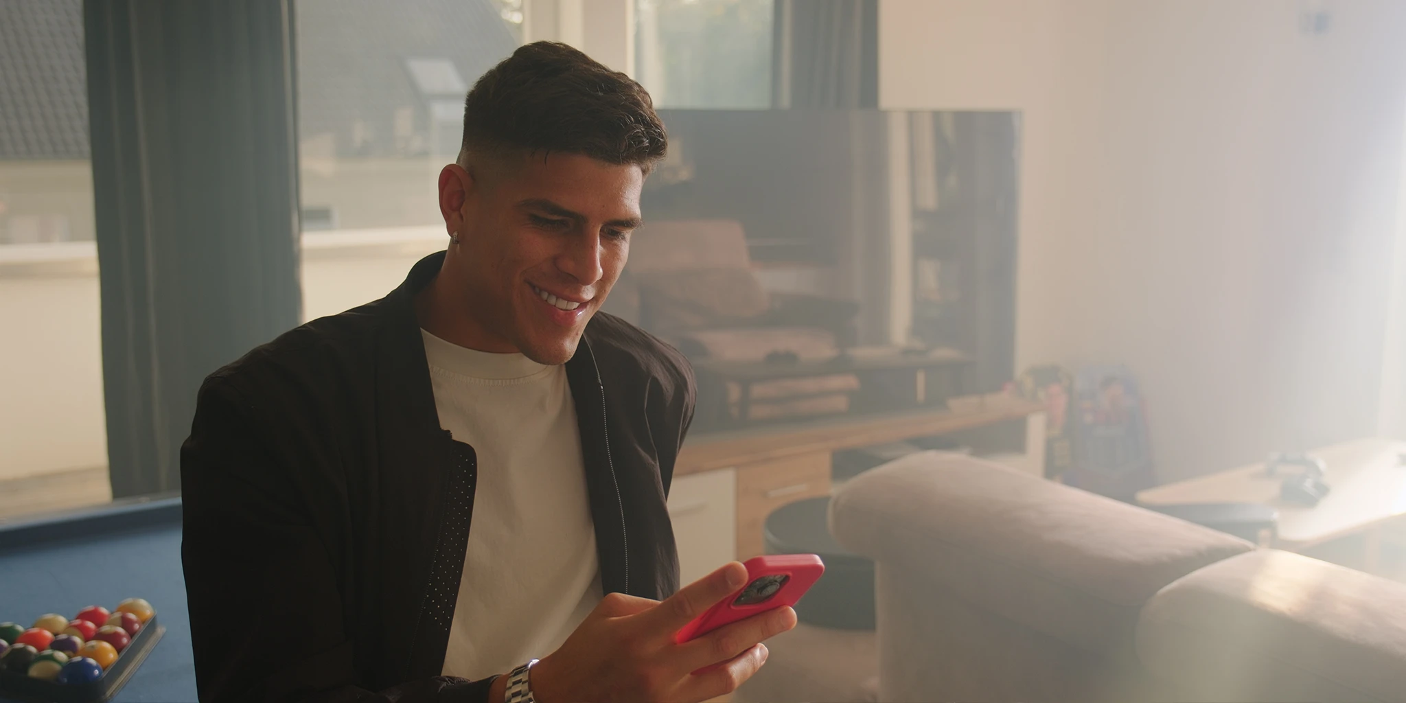 Soccer pro Piero (Piero Hincapié) sits in his villa on a pool table and reads a text message on his cell phone. He is wearing a plain black jacket and a white shirt. The low sun floods the room. A turned-off TV can be seen in the background.