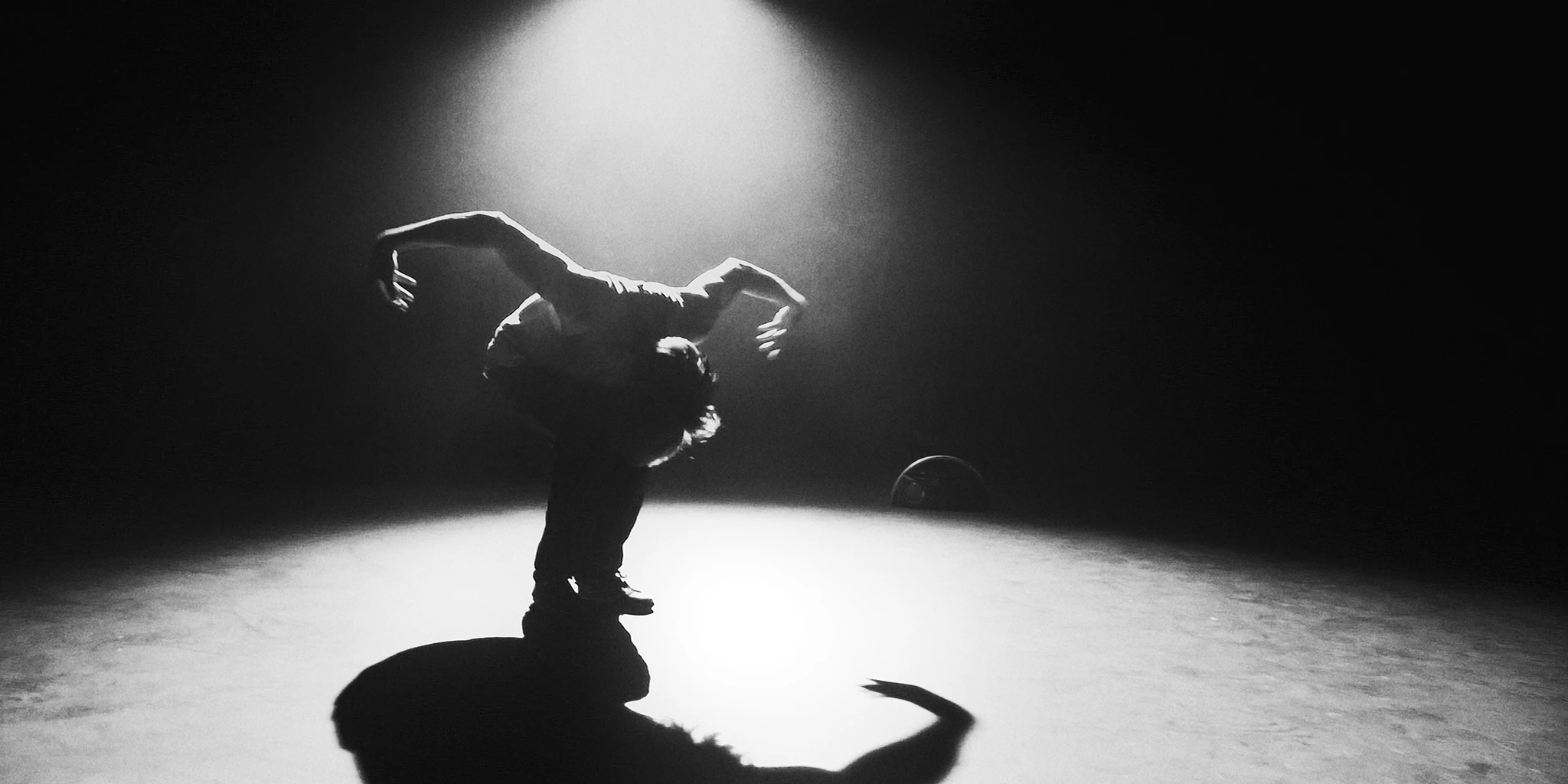 A young man dances in the middle of a spotlight. His torso is on his thighs and his knees are bent. His arms are outstretched like a swan. A Bluetooth speaker can be seen in the background.