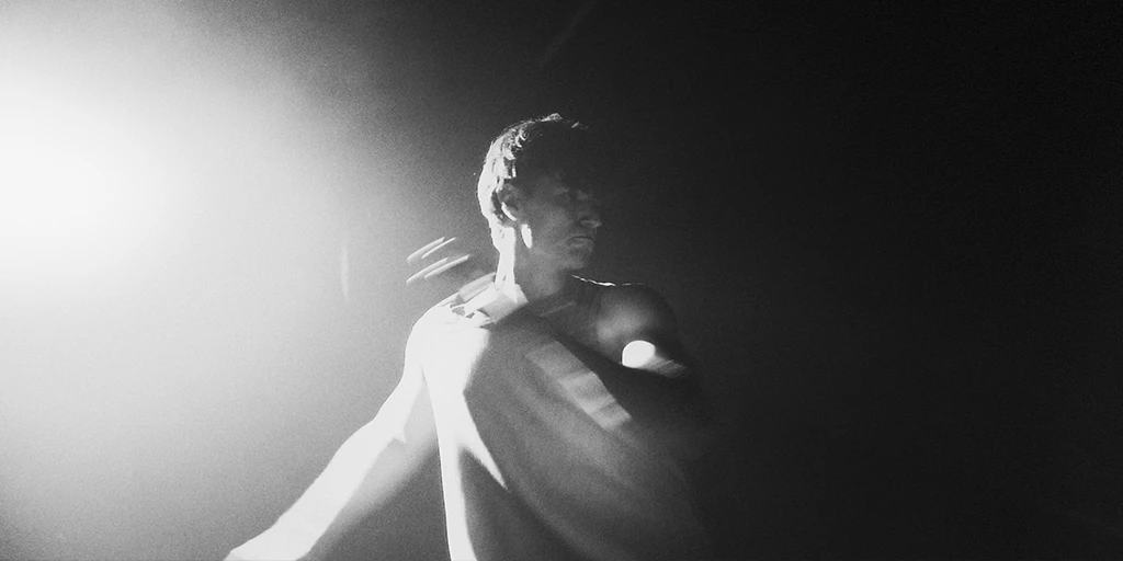 A young man with dark hair and a tank top looks to the right out of the picture. He comes from a dance step. Bright light hits his right side. The room behind him is dark.