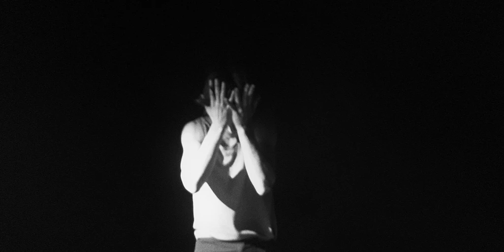 A young man presses his open palms against his forehead. He has black hair and is wearing a tank top. Bright light hits his right side.