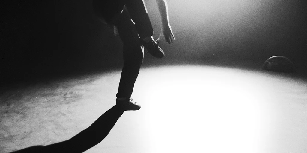 An oval white spotlight is seen on the floor. In the spotlight, a young man dances. His legs and left arm are visible. His right leg is bent. He is wearing black pants. A Bluetooth speaker stands in the background on the right.