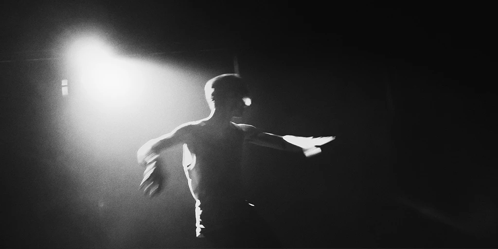 A young man spins in the bright light of a spotlight. He has dark hair, wears a gray tank top and black pants. The room is dimly visible in the background.