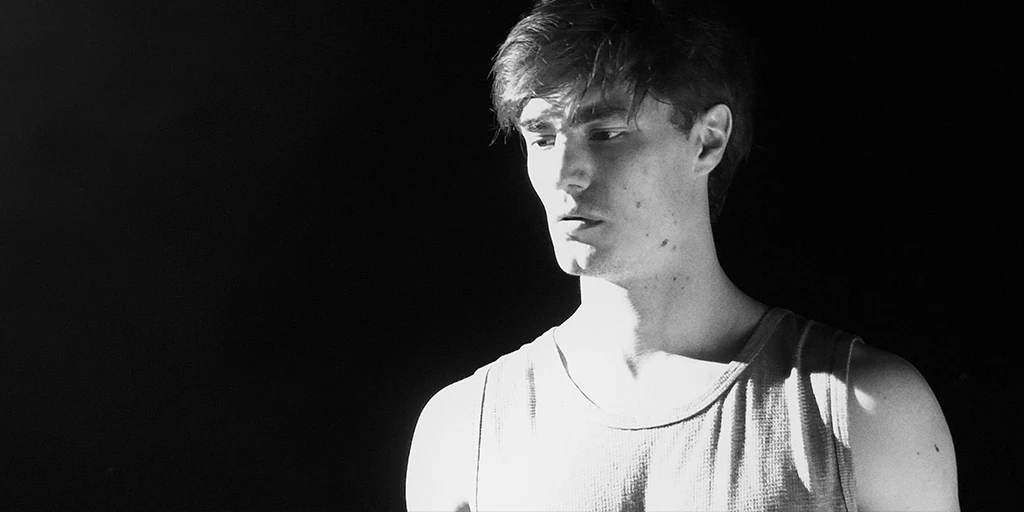 In a close-up, a young man is seen. He has dark hair and is wearing a tank top. His eyes seem to stare into space. The room behind him is dark.