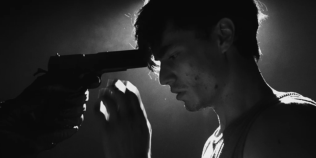 A young man with dark black hair is seen in profile. He reaches for the gun held to his head. Backlight hits the scene in the otherwise dark room.