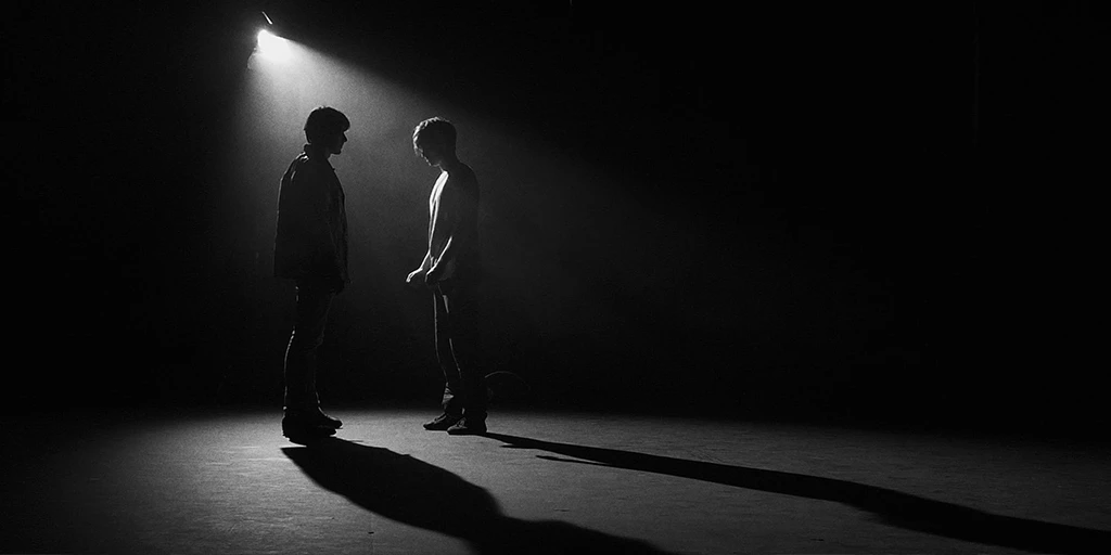 Two men stand facing each other. Backlight comes from a spotlight. The rest of the room is dark. Both stand at some distance and take a neutral, upright posture. A middle-aged man stands on the left and a young man stands on the right. The light reflects on the floor. Long shadows stretch through the room.