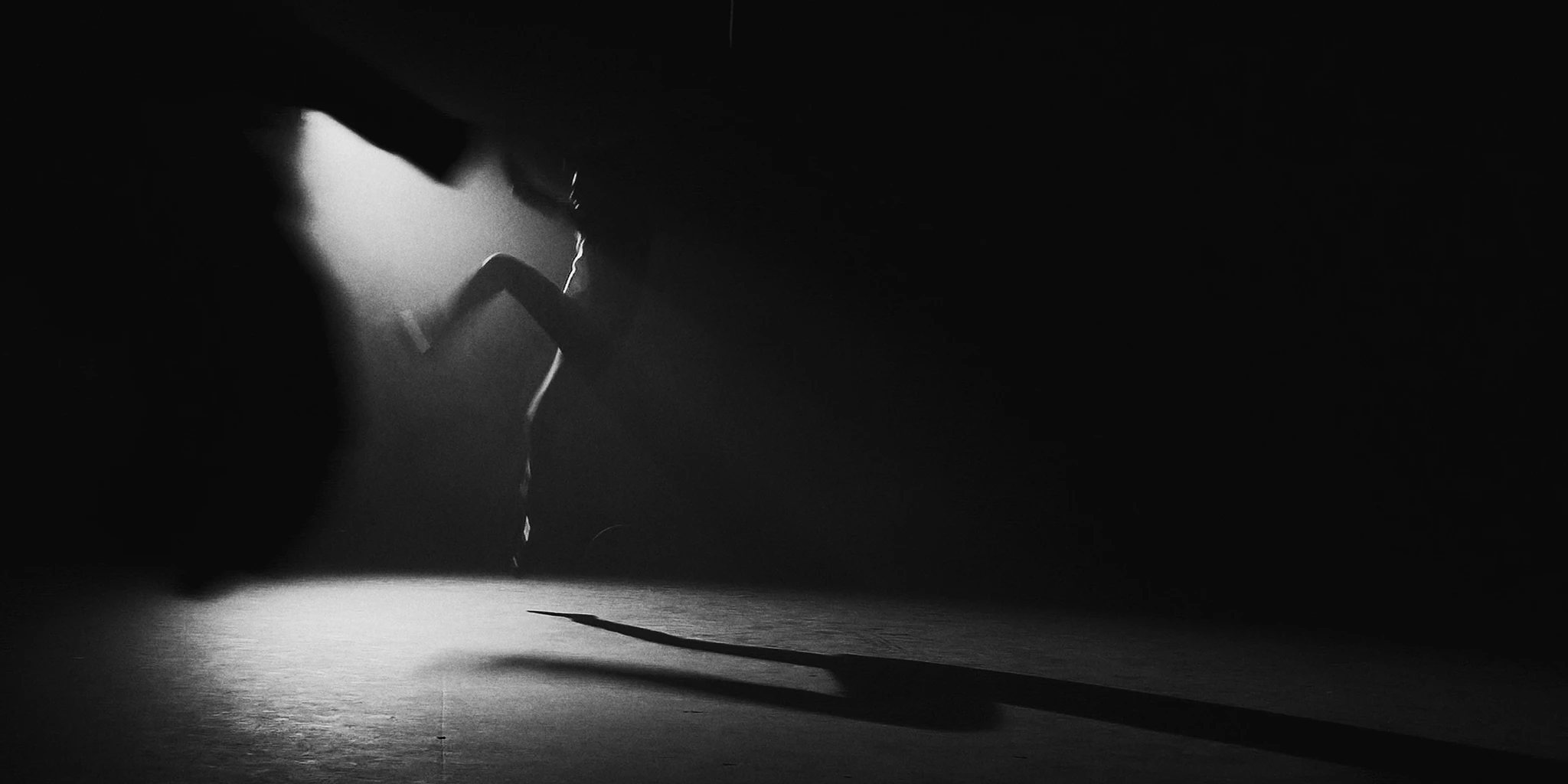 In the center of the image, a dancer is seen performing a jump. His right leg is slightly in the air, with his left leg he steps forward. His torso is slightly leaned back. In the background, there seems to be a light source that casts a spotlight on the floor of the otherwise dark room. The silhouette of a gun is visible in the left foreground. Graphically, it looks like the gun is aimed at the young man.