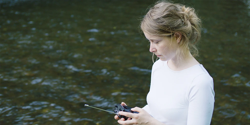 A woman, in her early 30s (Odine Johne), wades through a river. She operates a radio. She is wearing a white top and her blonde hair is tied back in the feature film »In the Sea of Silence«