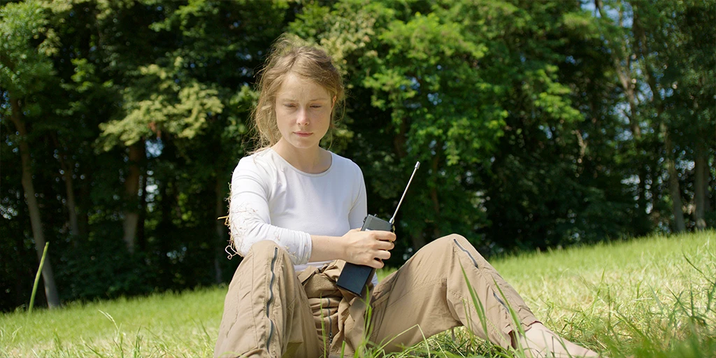 An astronaut (Odine Johne) holds a radio in her hand and looks into the grass. She is wearing a white top and brown pants in the feature film »In the Sea of Silence«