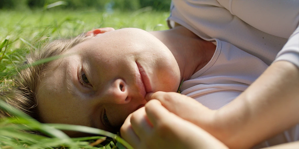 A young woman (Odine Johne) lies sideways in the grass and looks at something outside the frame. The sun hits her cheek. She is wearing a white top in the feature film »In the Sea of Silence« © Johannes Schmülling