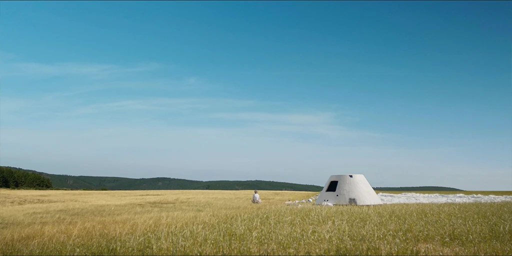 The astronaut (Odine Johne) sits next to her capsule and parachute in the grass, looking out into a grassy landscape. A forest is visible on the distant horizon in the feature film »In the Sea of Silence« © Johannes Schmülling