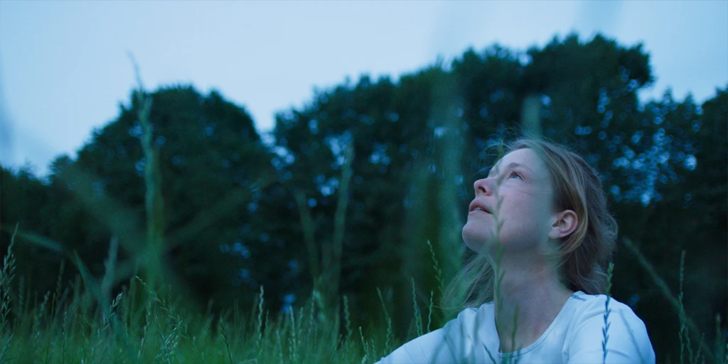 An astronaut (Odine Johne) sits in the grass and looks into the universe. Light wind blows through her hair in front of a bluish evening sky. She is wearing a white top in the feature film »In the Sea of Silence«