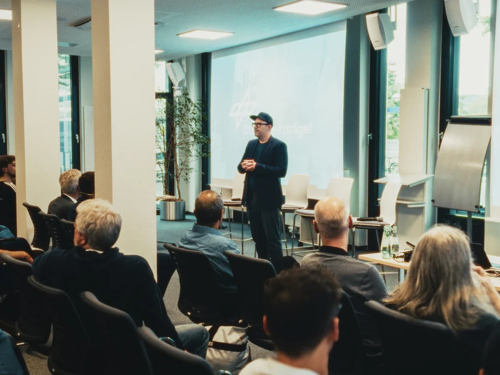 A conference room with a man in a cap and jacket standing on stage. The audience watches him.