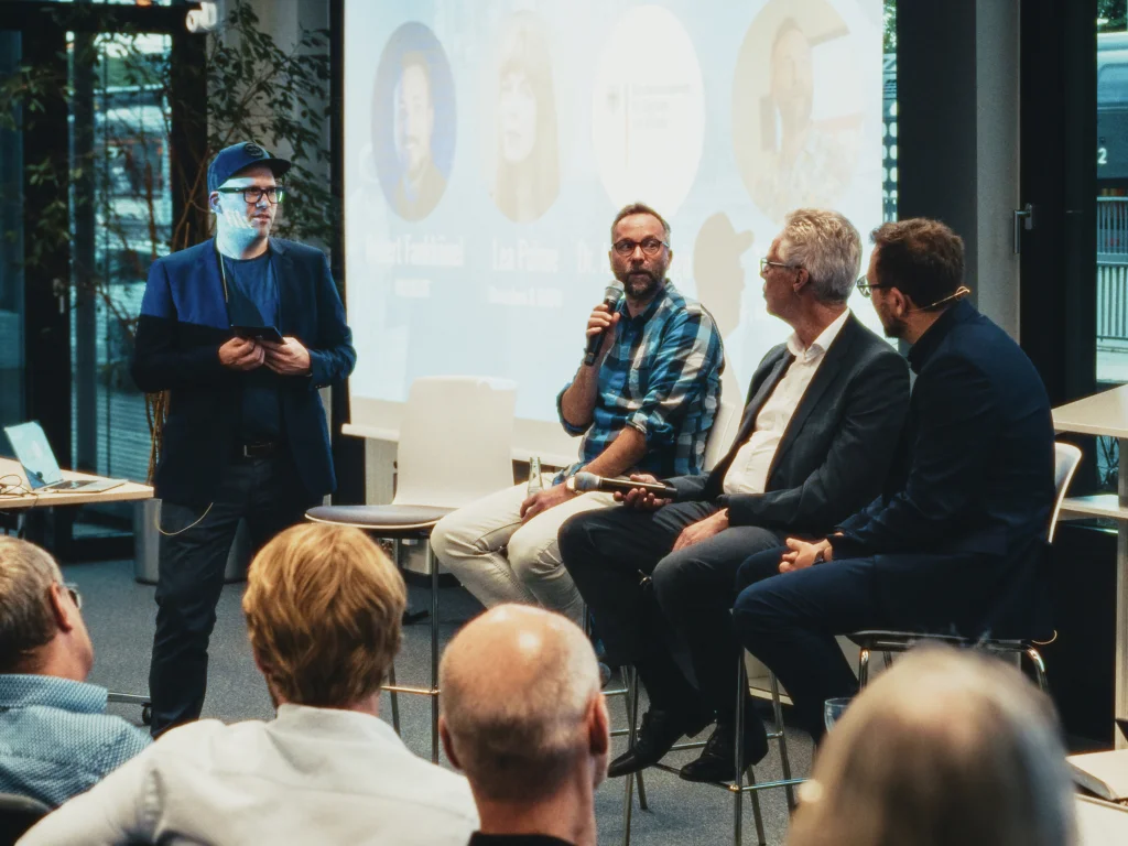 On the left is a man with a cap, on the right are three men sitting with a microphone in a conference room.