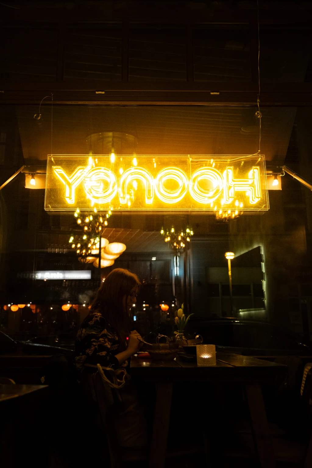 A woman sits in a restaurant under a yellow neon sign, eating.