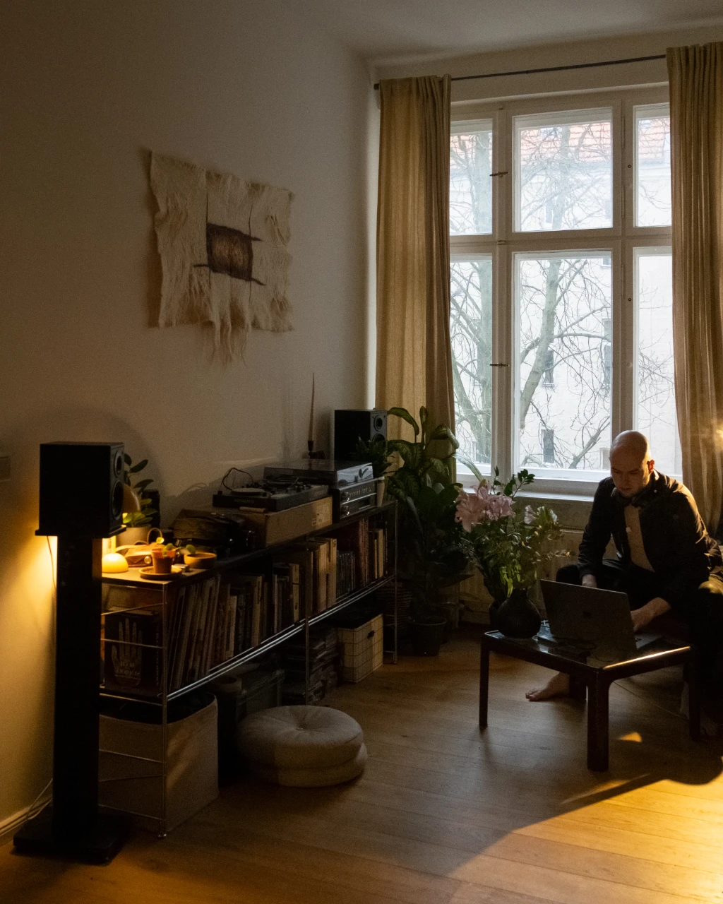 A young man sits in a large living room at his MacBook. Records are on the shelf to the left. Daylight comes through the window next to him.