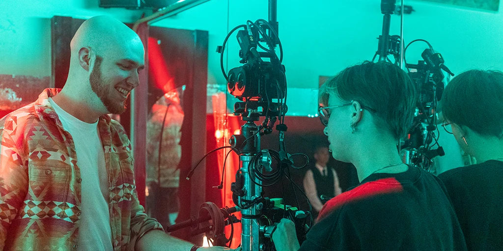 A young man and a young woman standing sideways in front of a mirror. In the middle, a Virtual Reality camera is placed. Both are working on the camera rig. The young man is smiling. The scene is bathed in red and green light.