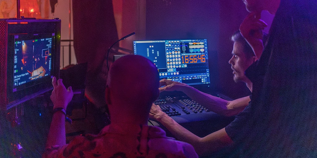 Three people in front of monitors. The young man on the left points to the monitor showing the image of the scene being filmed. Next to him on the right is a man following his instructions and operating a lighting console. Another man is visible on the edge of the frame. The monitors emit a bluish light.