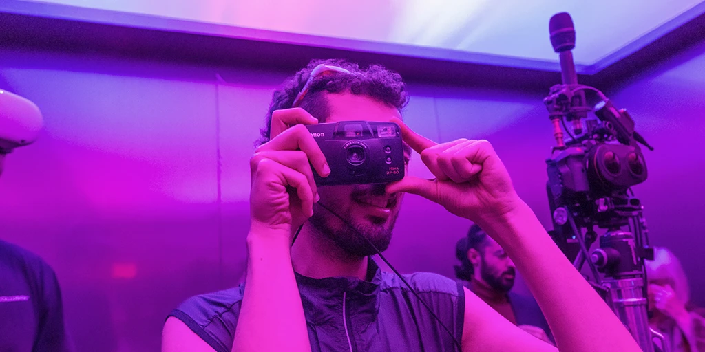 A man stands in an elevator taking a photo with an analog camera. Magenta and blue light shine through the transparent ceiling. To the right of the man is a Virtual Reality camera.