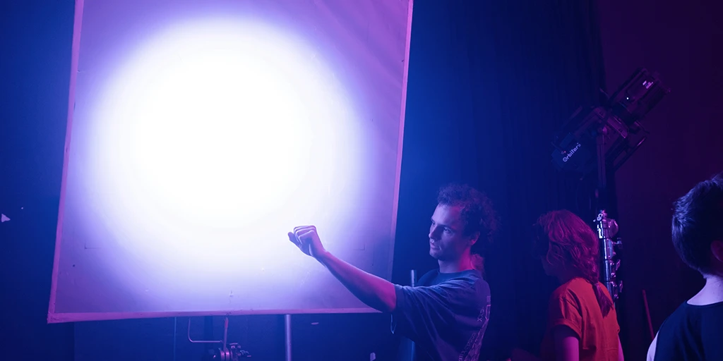 A young man stands in front of a 1x1 meter frame through which light is shining. He clenches his hand into a fist and checks the light. Next to him is a young woman in a red shirt. Bluish light floods the room.