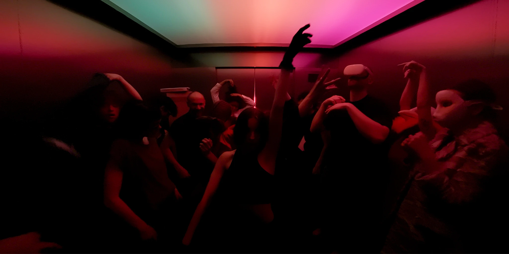 We are standing in an elevator, the ceiling light of which is bathed in a gradient of green, orange, and magenta. In front of us is a wild party crowd. A young woman stands directly opposite and raves by raising her arm. A tall man wears VR glasses. A woman standing on the right wears an animal mask. In total, there are about 10 people dancing to the techno beat.