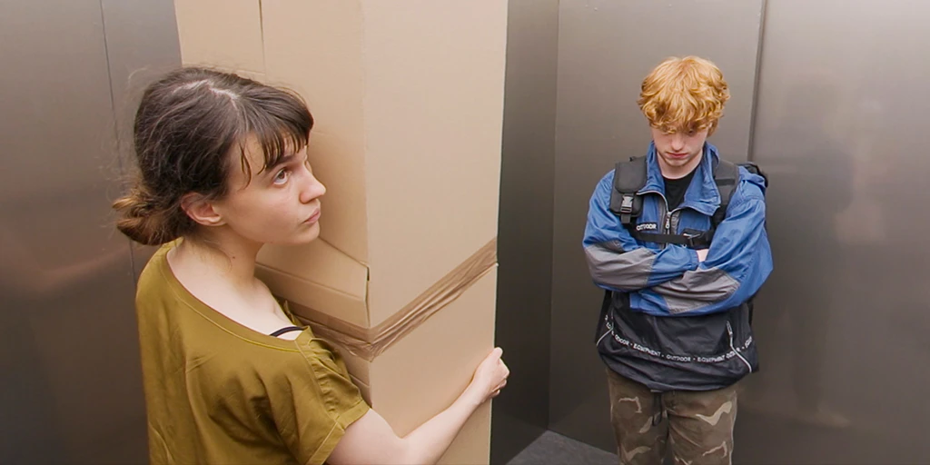 A young woman stands with a large box in an elevator and looks up at the ceiling. She has dark hair, brown eyes, and wears a yellow shirt. To her right stands a young man in a blue jacket. He has red hair, wears a backpack, and looks down.