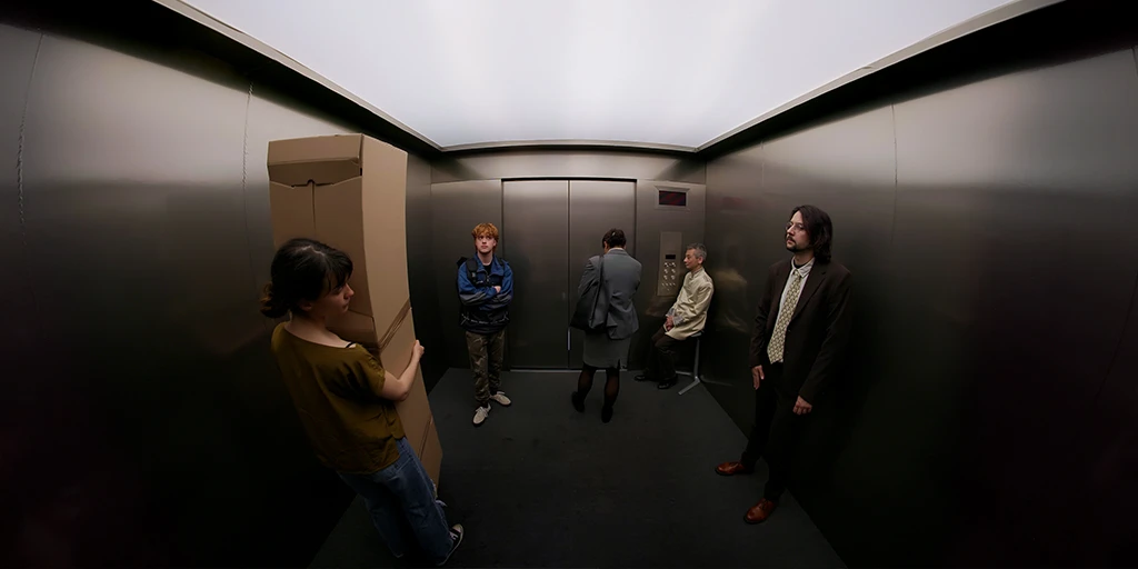 We are standing in a metal elevator. The ceiling seems to be made of frosted glass through which light falls. It seems somewhat dark in the elevator. On the left stands a young woman with a package, and to her right a man with a backpack. With their backs to us stands a woman who looks businesslike. To the right, a middle-aged man sits on a chair, controlling the elevator. In front of him stands a man with long hair. He wears a tie and a suit.