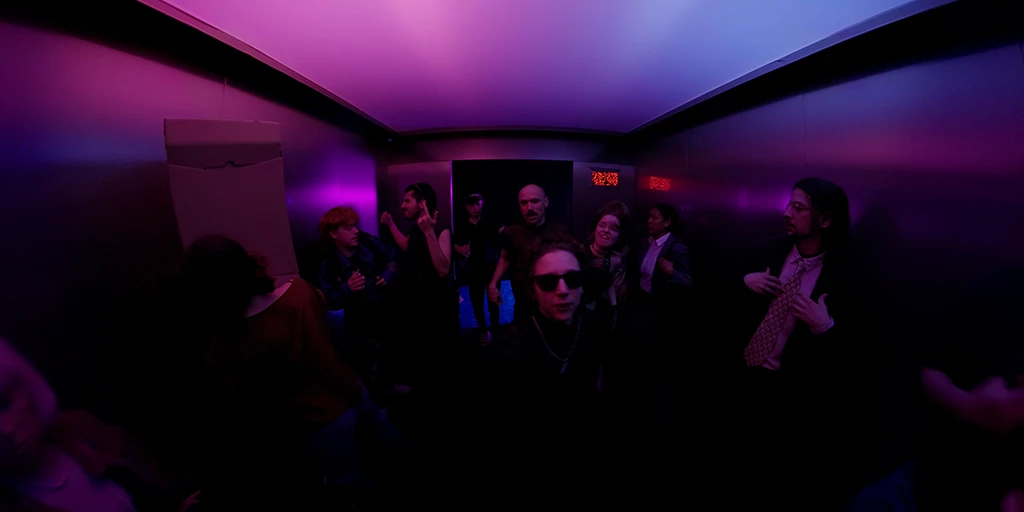 We are standing in a large elevator with the doors open. The outside world is dark. Through a large frosted glass ceiling, magenta-blue light shines into the elevator. Diversely dressed ravers enter the elevator. A small woman with short hair stands directly in front of us wearing dark sunglasses. The outlines of about 9 people can be seen.