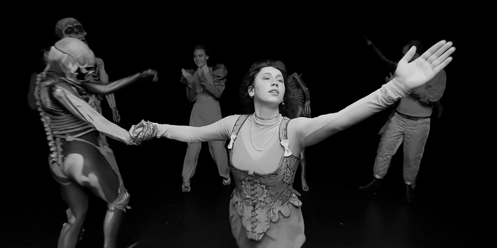 A pitch-black room. A woman with dark hair raises her arm and dances hand in hand with other people in skeleton costumes. She wears a traditional costume and a long necklace. In the background, a woman with light hair claps to the music. Three skeletons and three costumed people can be seen.