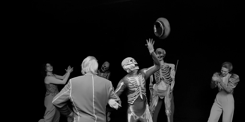 A dark room. Directly in front of us, an elderly man and a skeleton dance arm in arm in a circle. The skeleton has fun throwing a hat into the air. In the background, a woman claps her hands. Another person is in a skeleton costume. To the right, a skeleton puppet can be seen being controlled by a puppeteer. To the right of that, a young woman is seen clapping amusedly.