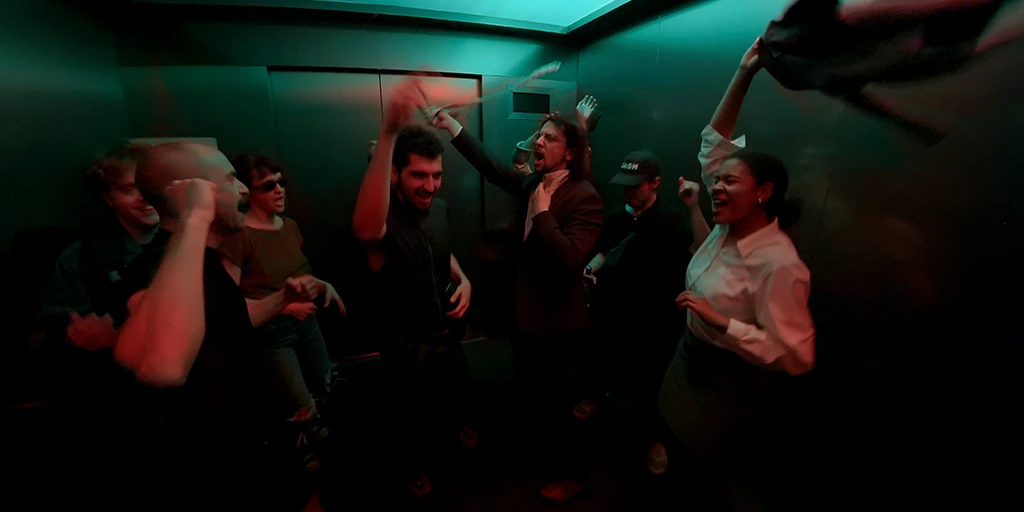 Eight people dance in a semicircle in a large elevator. Green-orange light hits them from above. A young woman in a business outfit throws a jacket through the elevator. A man in a suit waves his tie.