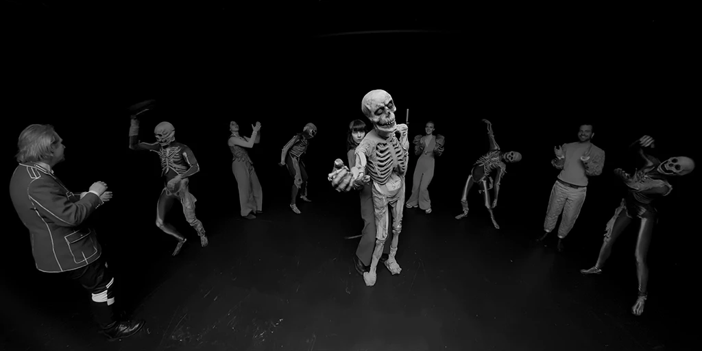 We are standing in front of a puppeteer with dark hair who is controlling a skeleton puppet and making an inviting gesture. Around her, a total of 8 people are dancing a dance of death. They are wearing skeleton costumes among other things.