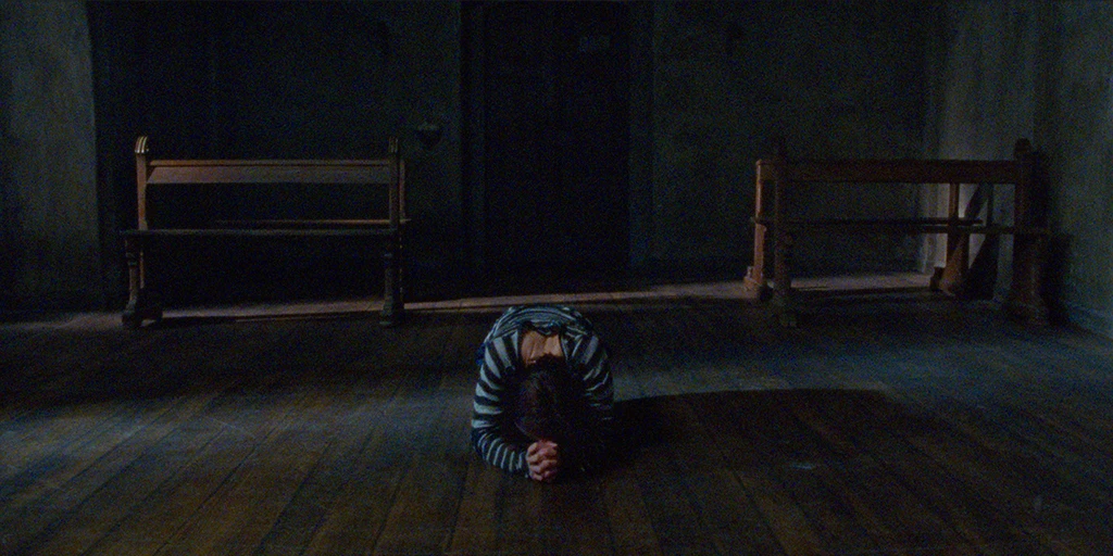 Leaning forward, the young woman kneels on the chapel floor. Behind her are two church pews. Daylight shines into the chapel from the side.