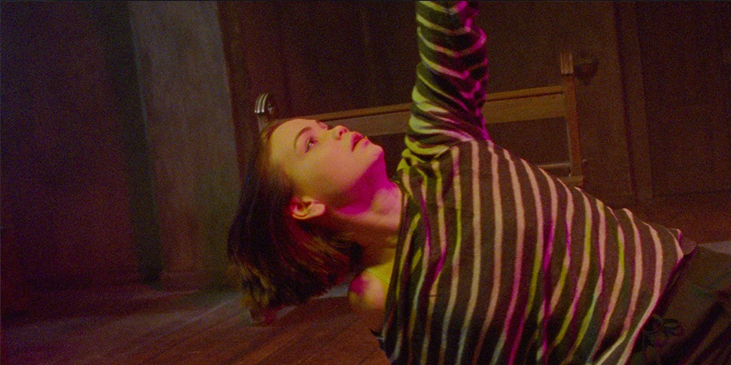 A young woman (Tatiana Feldman) moves in a dance pose in a chapel. Her left arm is stretched upwards. Yellow-magenta light hits her face.