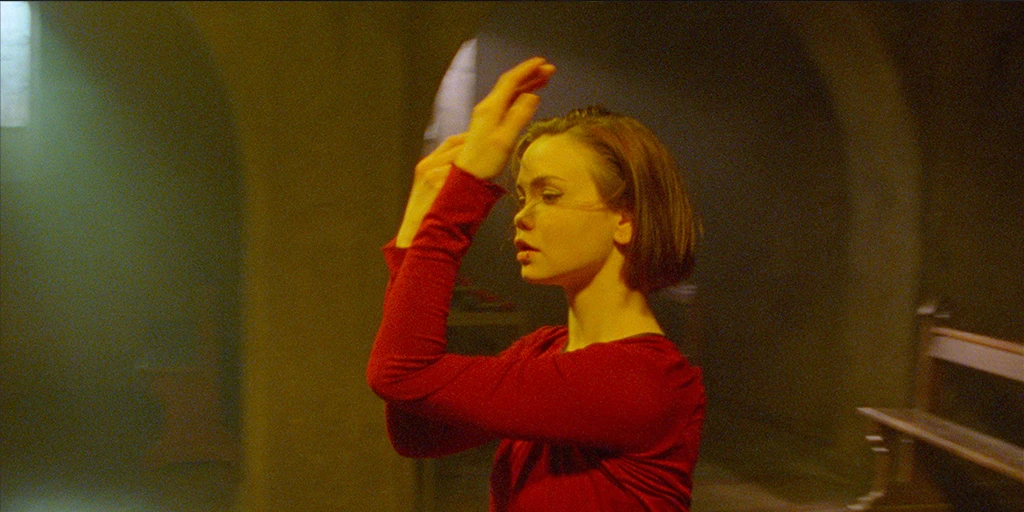 A young woman (Tatiana Feldman) dances in a chapel. She holds both hands in front of her face. Colorful light hits her face. In the background, daylight shines through the chapel windows.