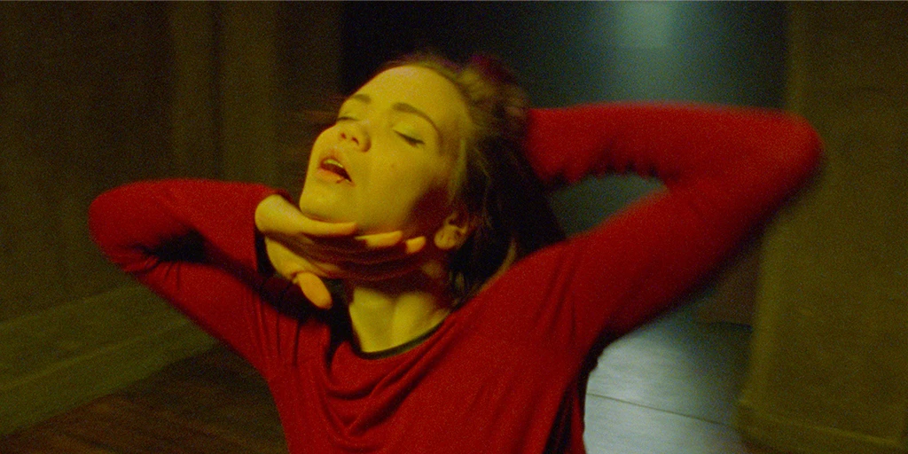 A young woman (Tatiana Feldman) dances in a red dress in a chapel. Her right hand brushes under her chin, her left hand is behind her head.