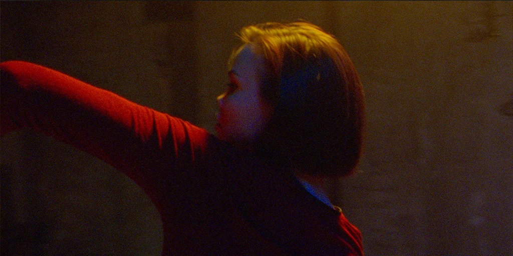 A believer (Tatiana Feldman) dances in a chapel. She is wearing a red dress. She stretches her left arm upwards.