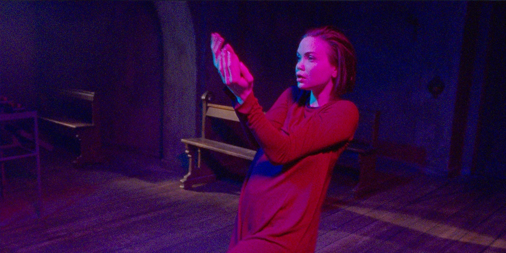 The believer (Tatiana Feldman) dances leaning slightly back. Magenta light hits her face, and she is wearing a red dress. Her hair falls evenly backward.