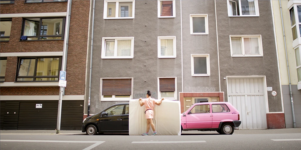 The protagonist of the film »The Mathematics of Things« transports a mattress. She wears a pink dress and walks past a pink car. In the background is the house of the man who gave her the mattress.