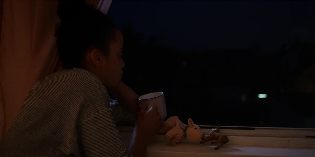 A young woman (Shari Asha Crosson) sits at an open window with a cup of tea at night. In front of her are two stuffed animals. She looks outside.