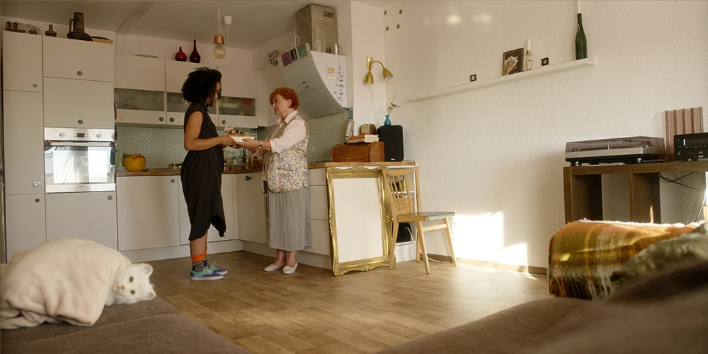 An elderly lady (Margit Laue) hands a box of stuffed animals to the protagonist Lena (Shari Asha Crosson) of the film »The Mathematics of Things« in her apartment. A cat lies on the sofa and stares at the camera. The room is flooded with sunlight.