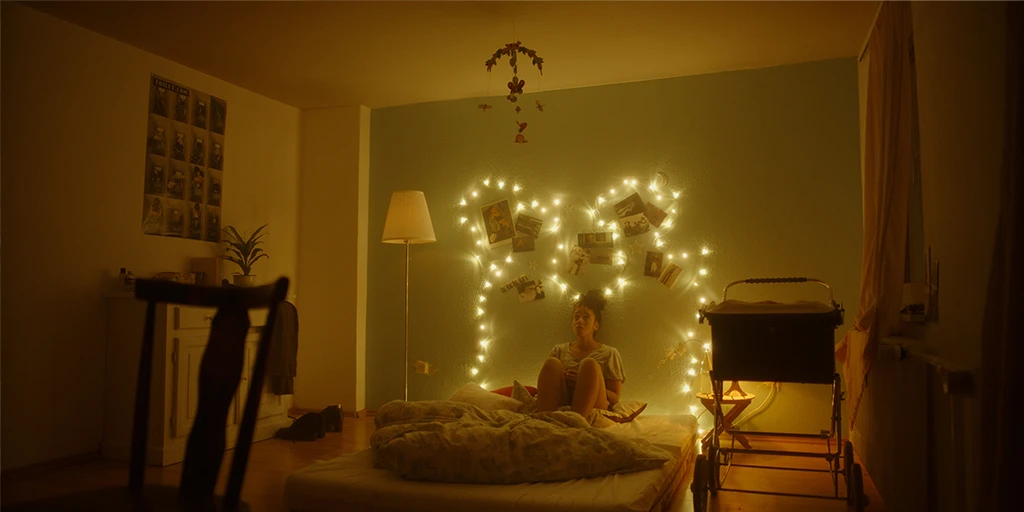 A young woman, Lena (Shari Asha Crosson) sits in a nursery. She looks at a mobile. Behind her, a string of lights shines. Lena looks thoughtful. The room is bathed in warm light.