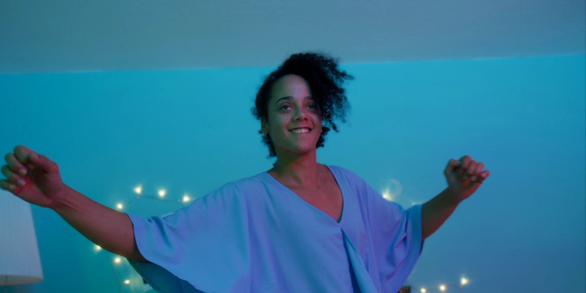 A young woman (Shari Asha Crosson) dances openly and exuberantly in front of a blue-green wall. A string of lights is in the background.